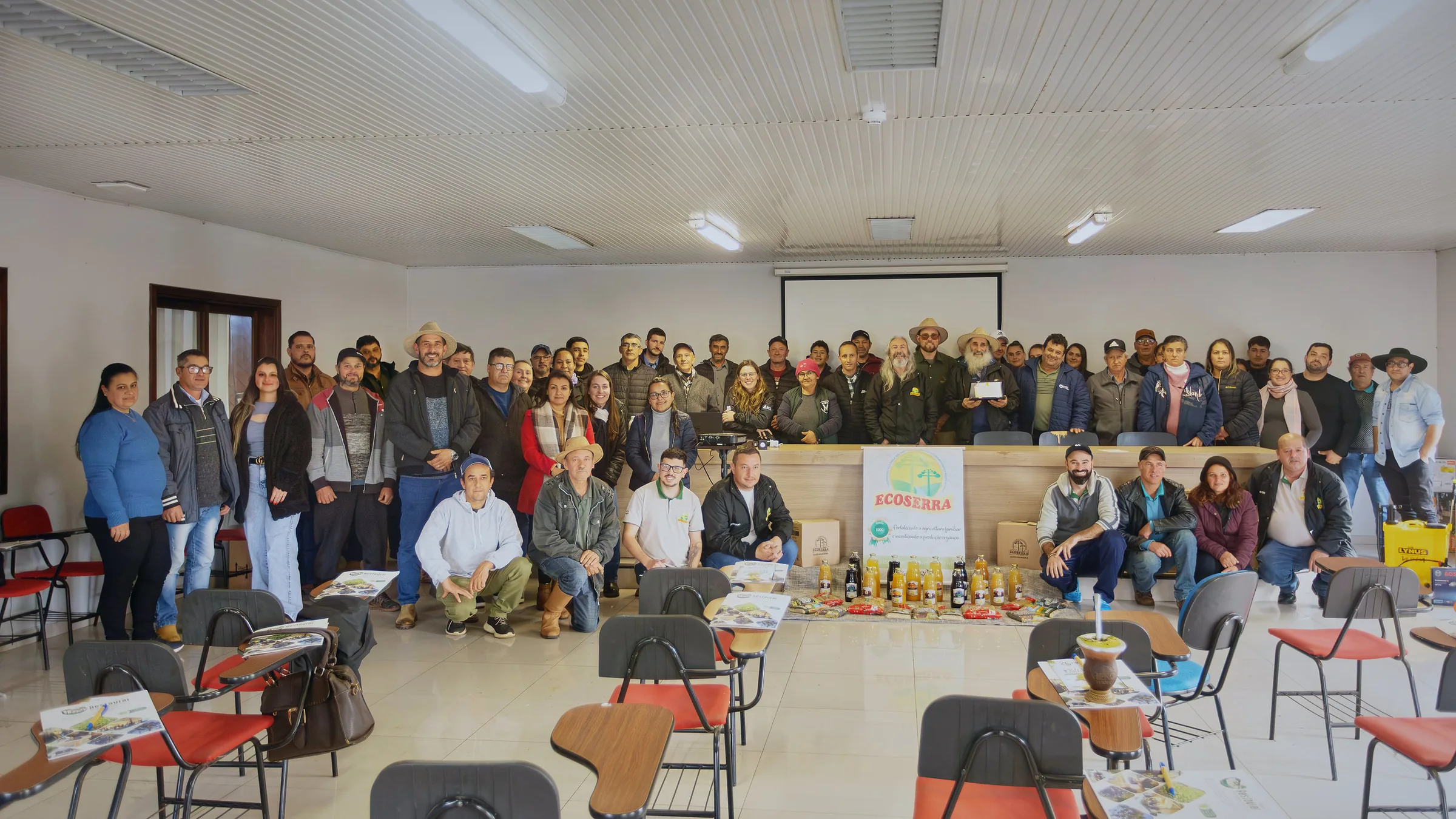 Agricultores e agricultoras cooperados da Ecoserra reunidos num auditório, com o banner da cooperativa e os produtos das famílias à frente